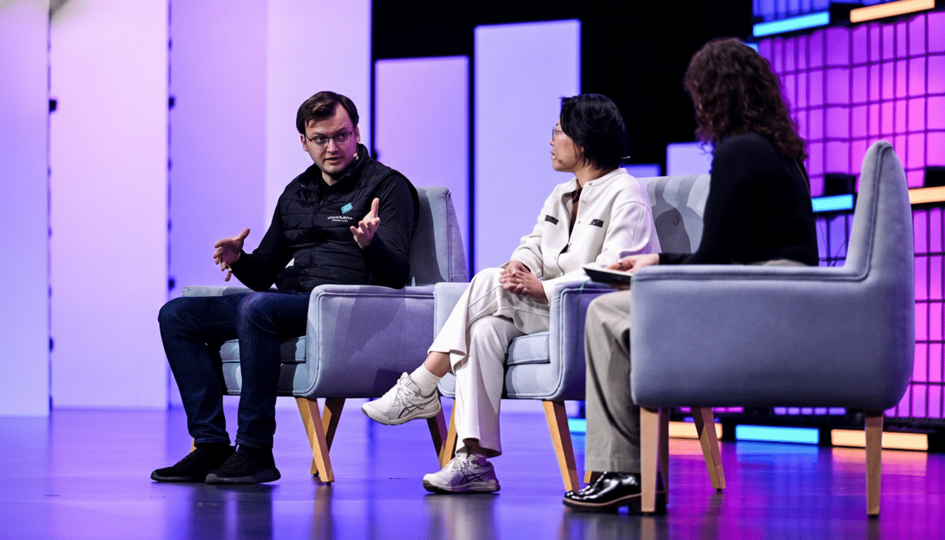 Three people are seated on stage in armchairs, engaged in a discussion. The man on the left is speaking with animated hand gestures, while the woman in the middle looks towards him. The third person on the right is partially visible from the back. The stage has a modern background with purple and white illuminated panels.