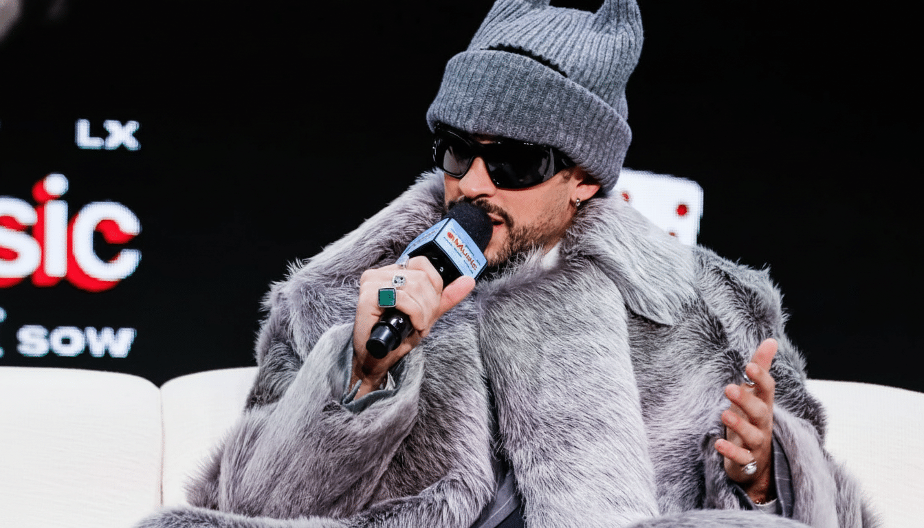 A man wearing a grey fur coat, sunglasses, and a grey beanie with cat ears, speaking into a microphone.