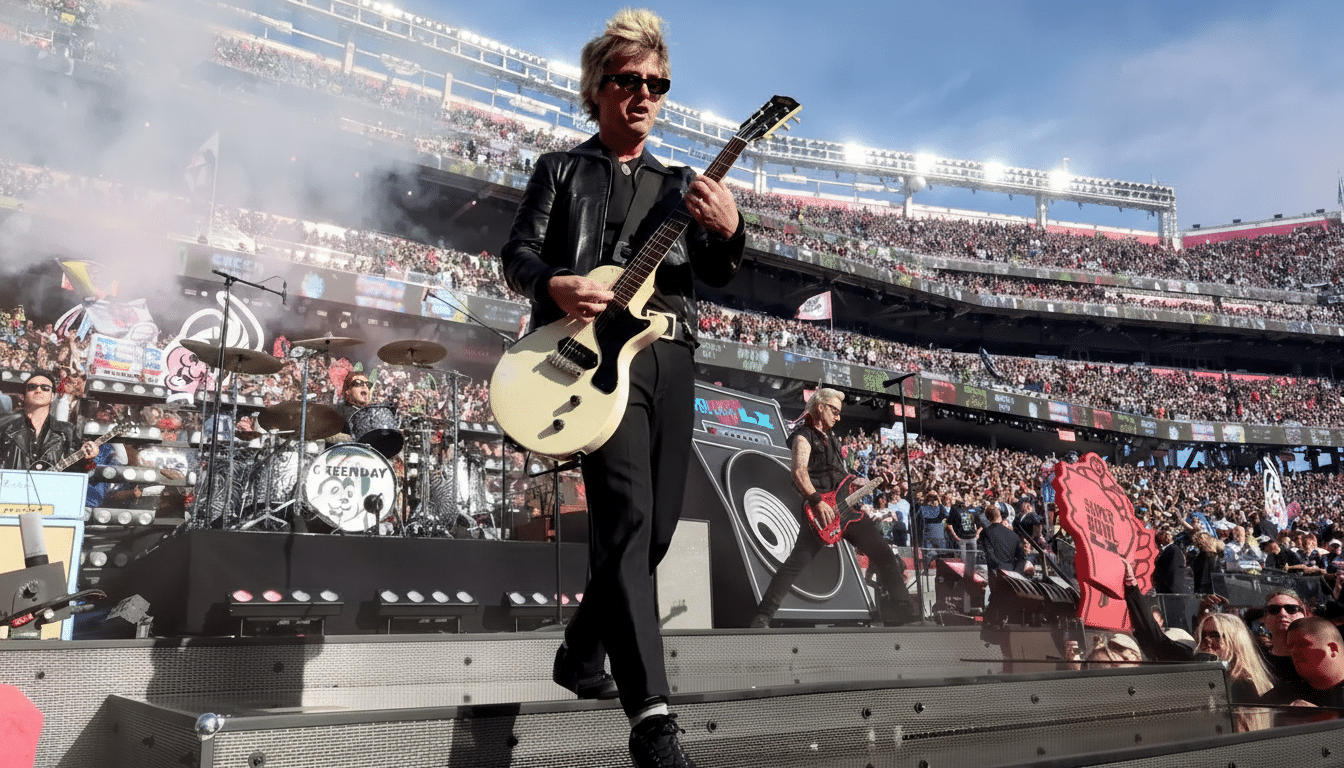 A musician in a black leather jacket and sunglasses plays a white electric guitar on stage at a large outdoor concert, with a drummer and another guitarist visible behind him, and a stadium full of cheering fans in the background under a clear sky.