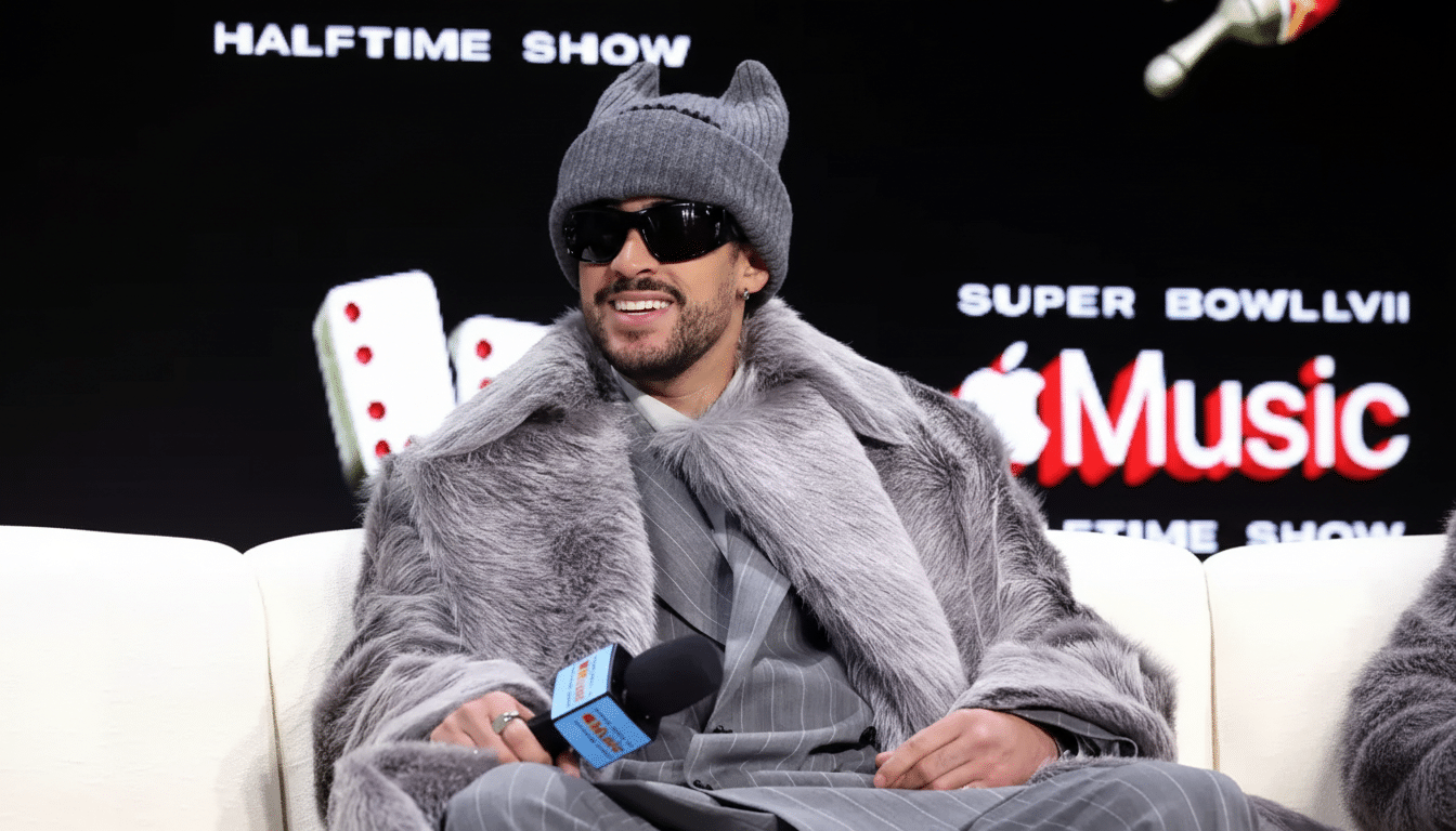 A man wearing a grey fur coat, sunglasses, and a grey beanie with cat ears, sitting on a white couch in front of a black background with HALFTIME SHOW and Apple Music logos.