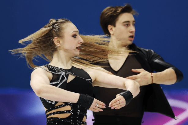 A female ice dancer with long blonde hair flowing to her right, wearing a black costume with cutouts and embellishments, is in the foreground. Her male partner, also in black, is slightly out of focus behind her.
