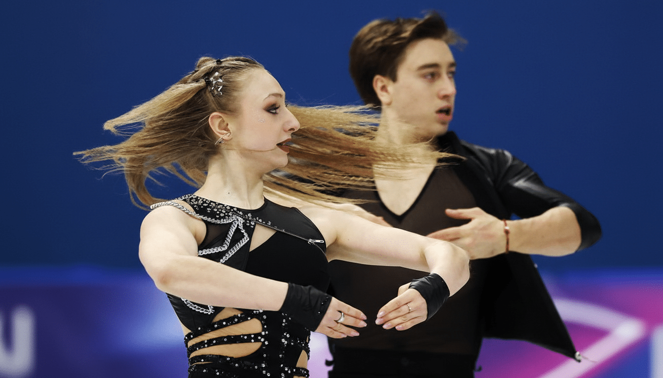 A female ice dancer with long blonde hair flowing to her right, wearing a black costume with cutouts and embellishments, is in the foreground. Her male partner, also in black, is slightly out of focus behind her.