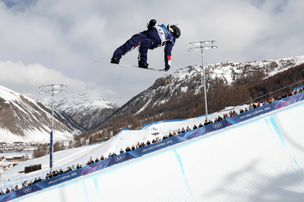 A snowboarder in a blue and red suit is captured mid-air against a backdrop of snow-covered mountains and a cloudy sky, with spectators visible on the slope below.