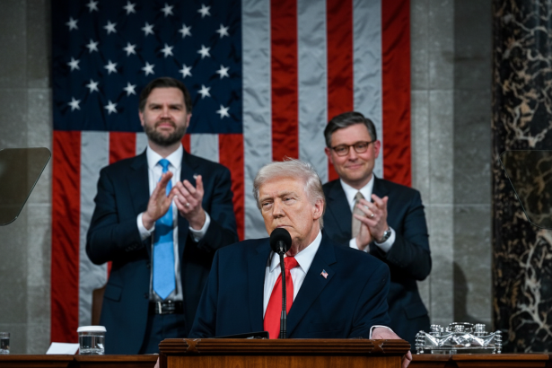 Donald Trump speaking at a podium with two men clapping in the background, in front of an American flag.