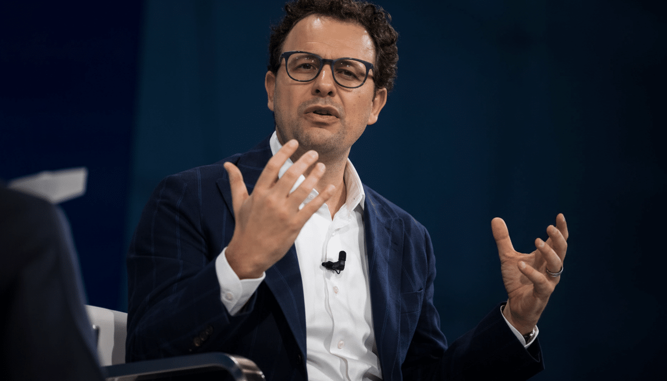 A man with dark curly hair and glasses, wearing a navy suit jacket and white shirt, speaking with his hands raised.