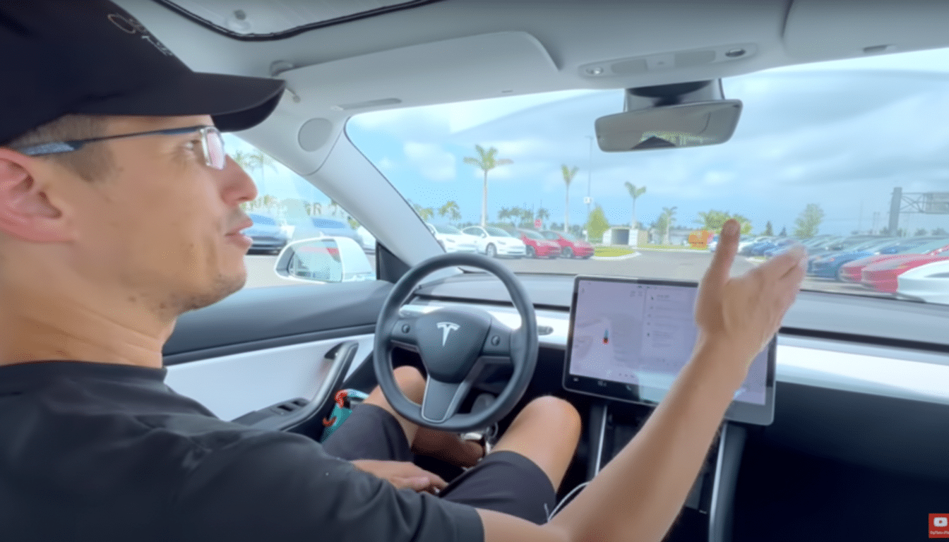 A man in a black cap and glasses is seated in the drivers seat of a white Tesla, gesturing with his left hand towards the right side of the car. The cars interior is visible, including the steering wheel with the Tesla logo and the large central touchscreen display. Through the windshield, a car dealership lot with several parked cars and palm trees under a cloudy sky can be seen.
