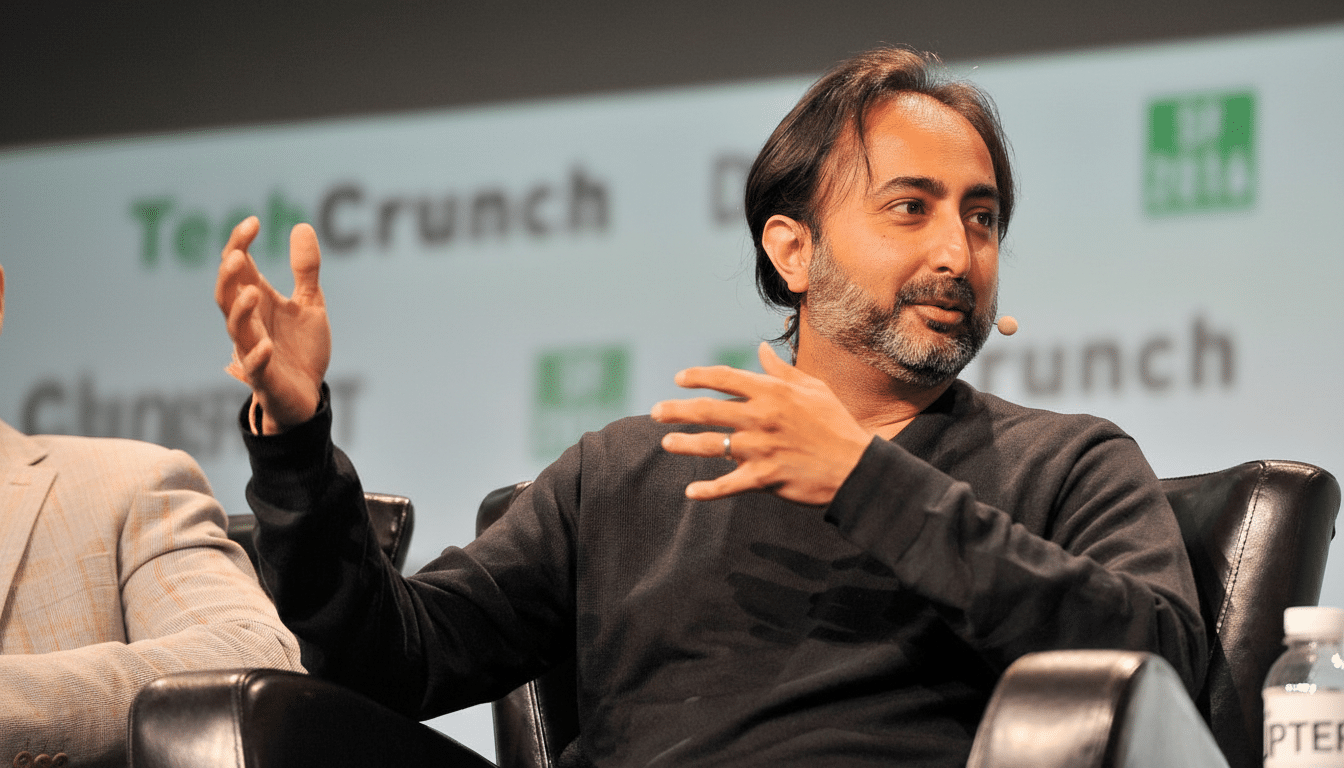 A man with dark hair and a beard, wearing a black sweater, sitting in a black leather chair and gesturing with both hands while speaking at a TechCrunch event.
