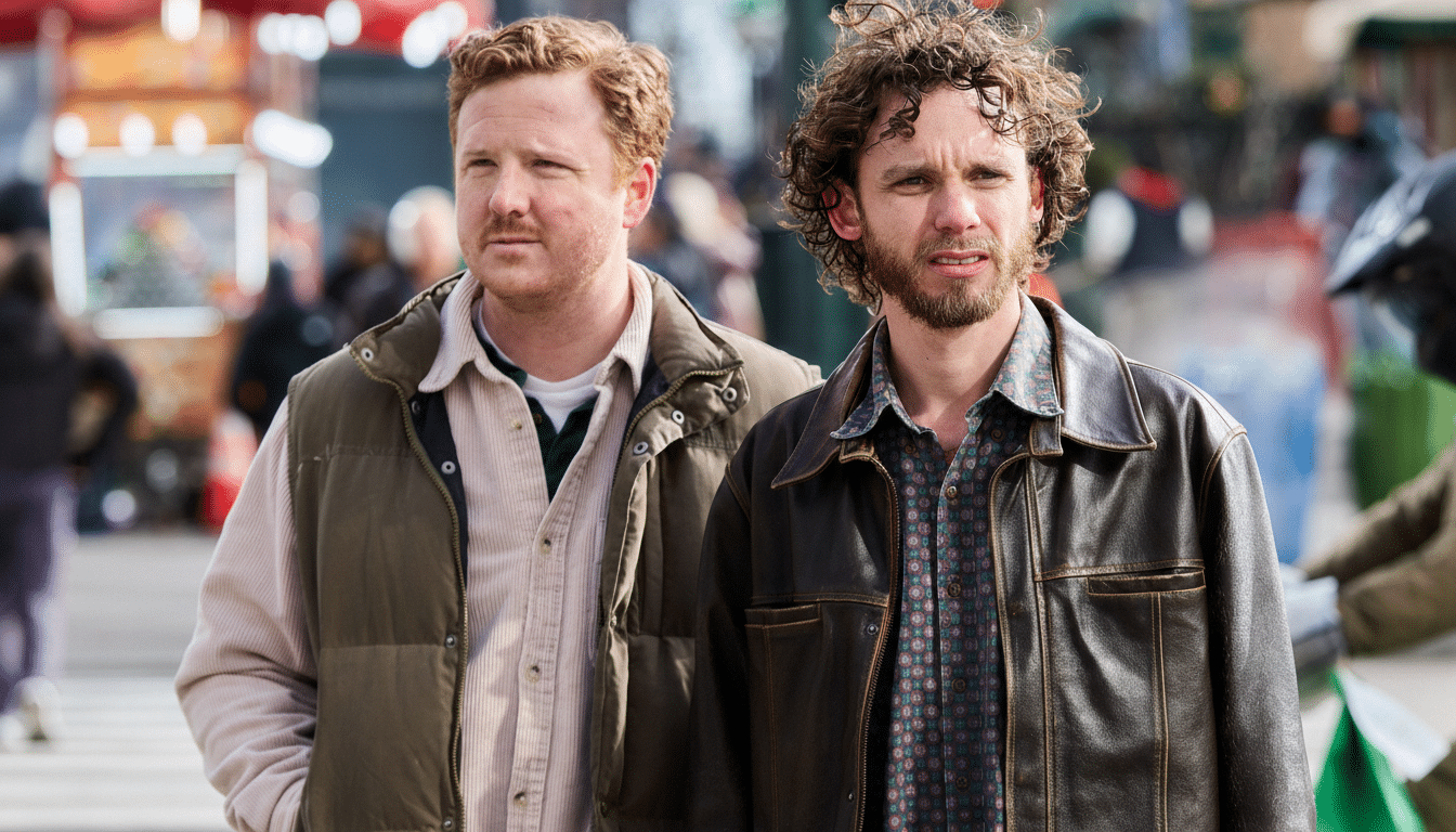 Two men, one with red hair and a vest, and the other with curly brown hair and a leather jacket, stand outdoors with a blurred background.