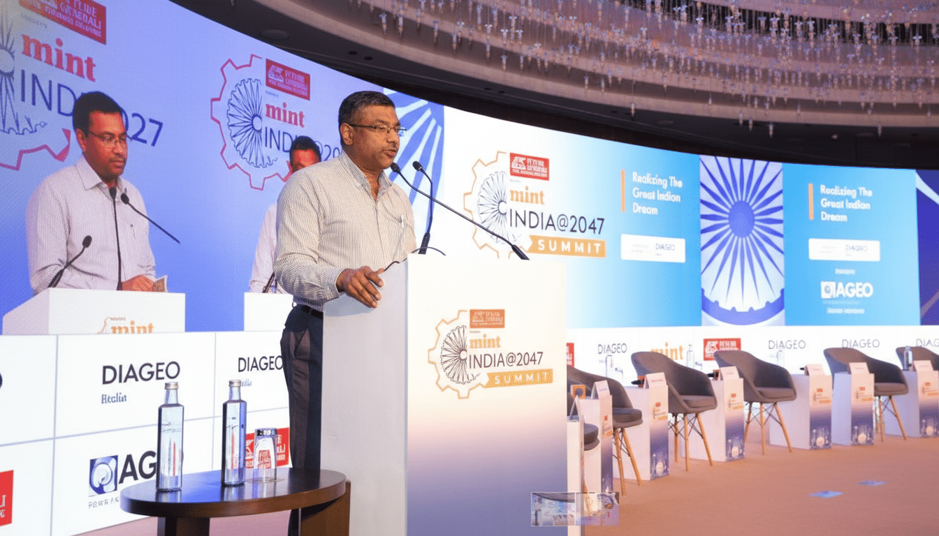 A man in a striped shirt speaking at a podium with mint INDIA@2047 SUMMIT branding, with a large screen in the background displaying event details.