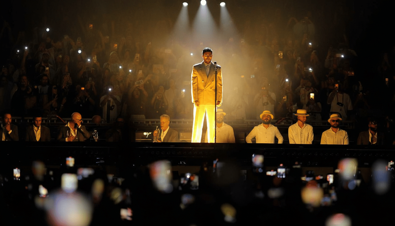 A male performer in a light-colored suit stands center stage under a spotlight, facing a large audience holding up their phones with lights on. Behind him, several musicians are seated.