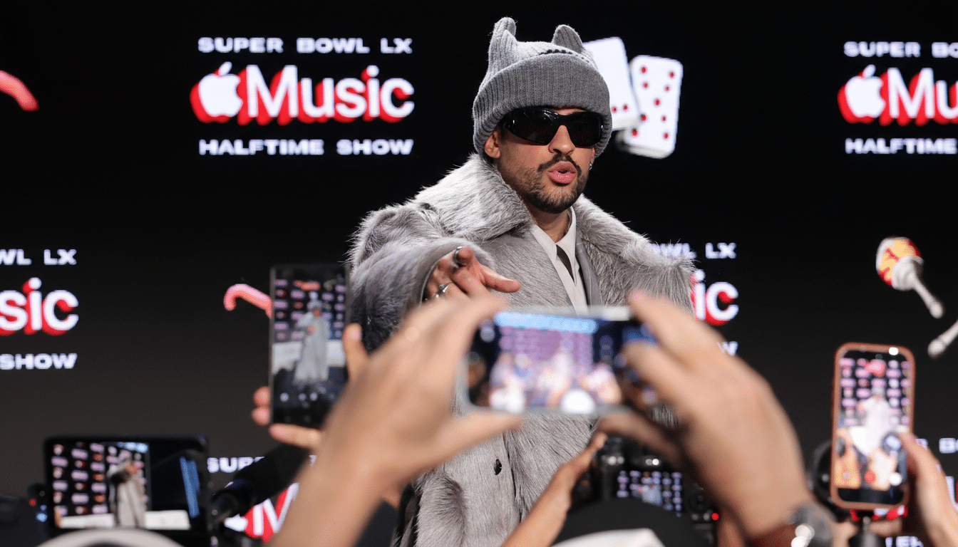 A man in a grey fur coat, sunglasses, and a grey beanie with ears stands in front of a black background with SUPER BOWL LX Music HALFTIME SHOW and an Apple logo. Several hands in the foreground hold up smartphones, seemingly recording him.