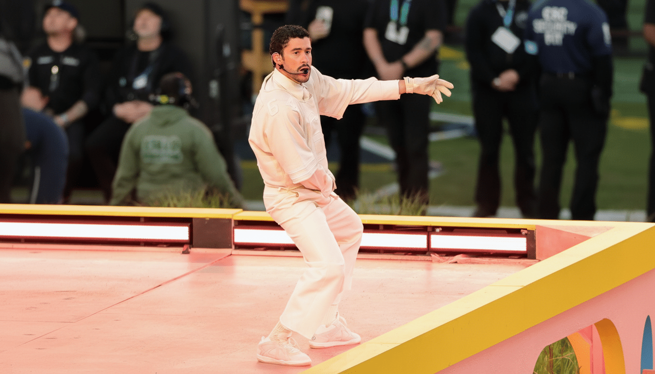 A man in a light-colored outfit performing on a stage with a microphone headset, extending his arm to the side.