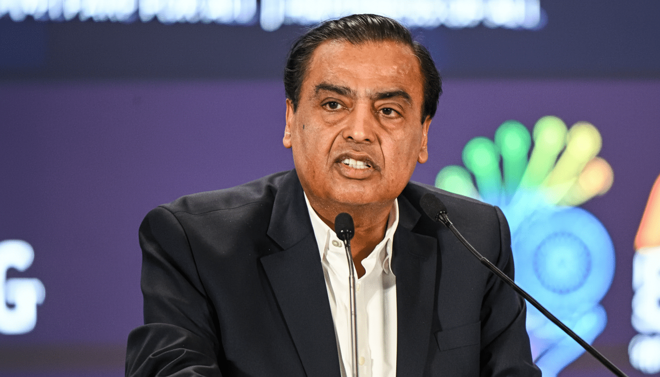 A man in a dark suit and white shirt speaking into two microphones, with a blurred background featuring colorful graphics.