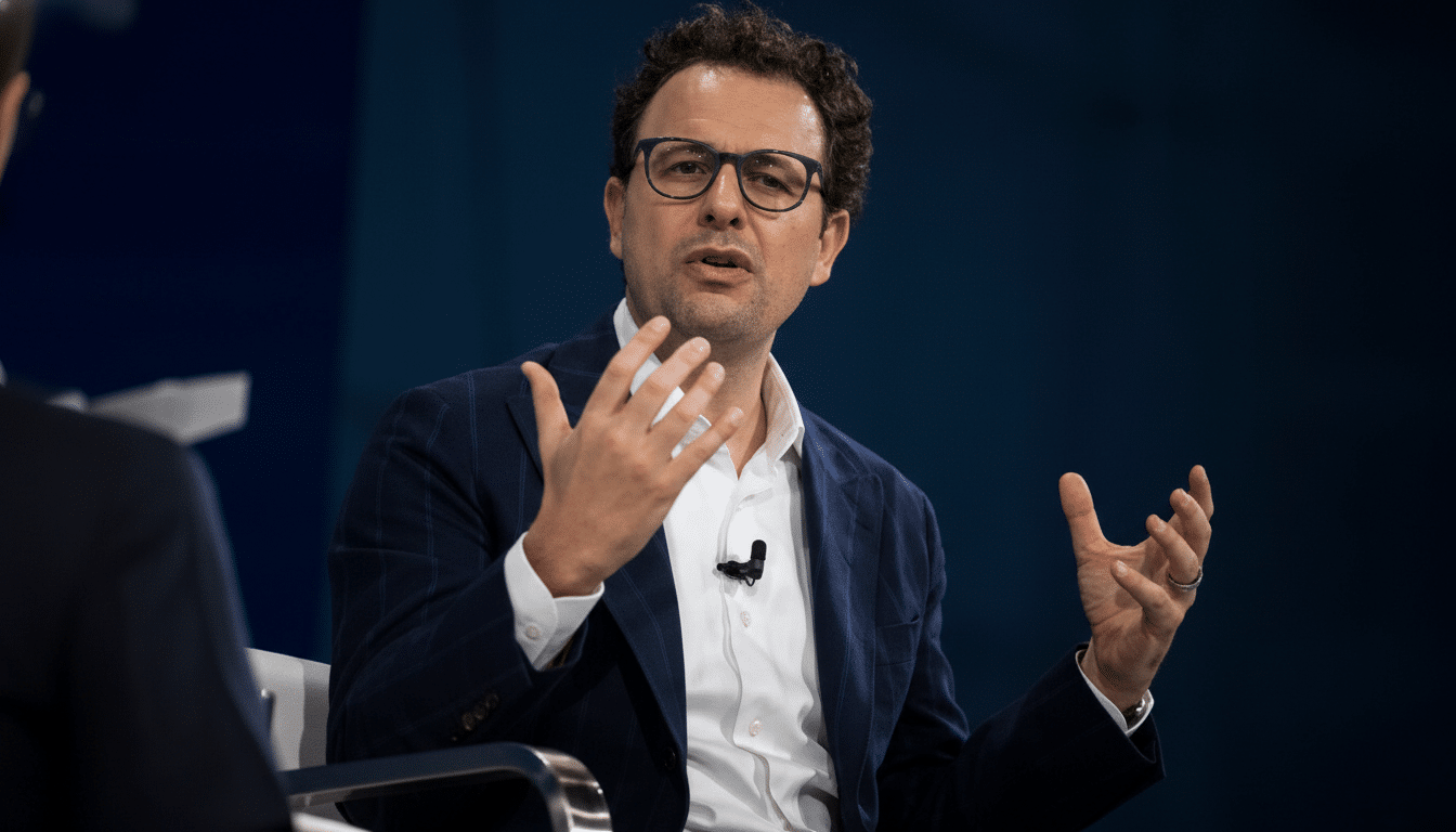 A man in a suit and glasses speaking with his hands gesturing, resized to a 16:9 aspect ratio.