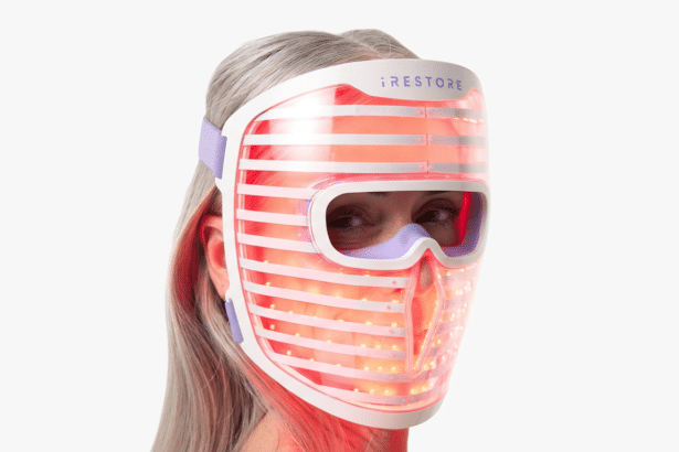 A woman with gray hair wearing an iRESTORE light therapy mask with glowing red lights, looking to the side.
