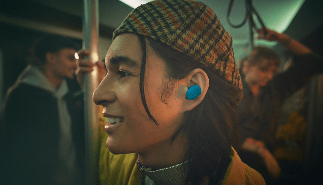 A young person with long dark hair and a plaid cap smiles while wearing a blue JBL earbud in their right ear, in a 16:9 aspect ratio.