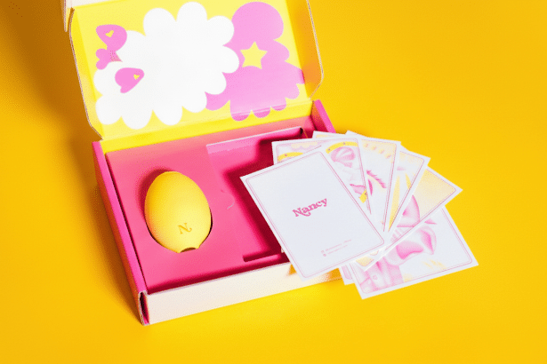 A yellow egg-shaped device with the letter N on it, nestled in a pink box alongside a stack of cards, all set against a vibrant yellow background.