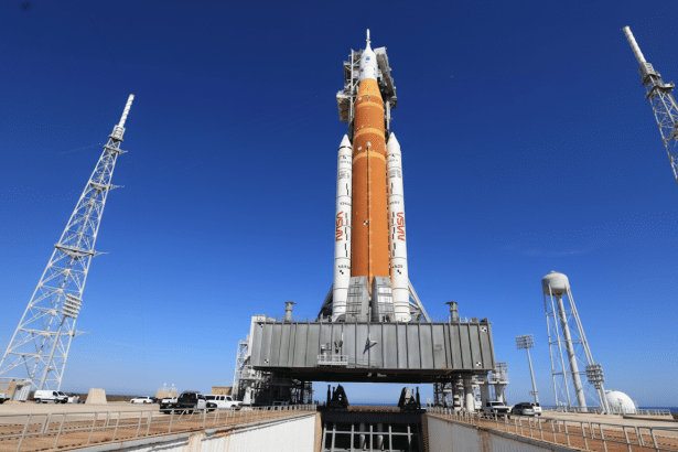 A large, orange and white rocket stands tall on a launchpad under a clear blue sky, flanked by two tall metal towers.