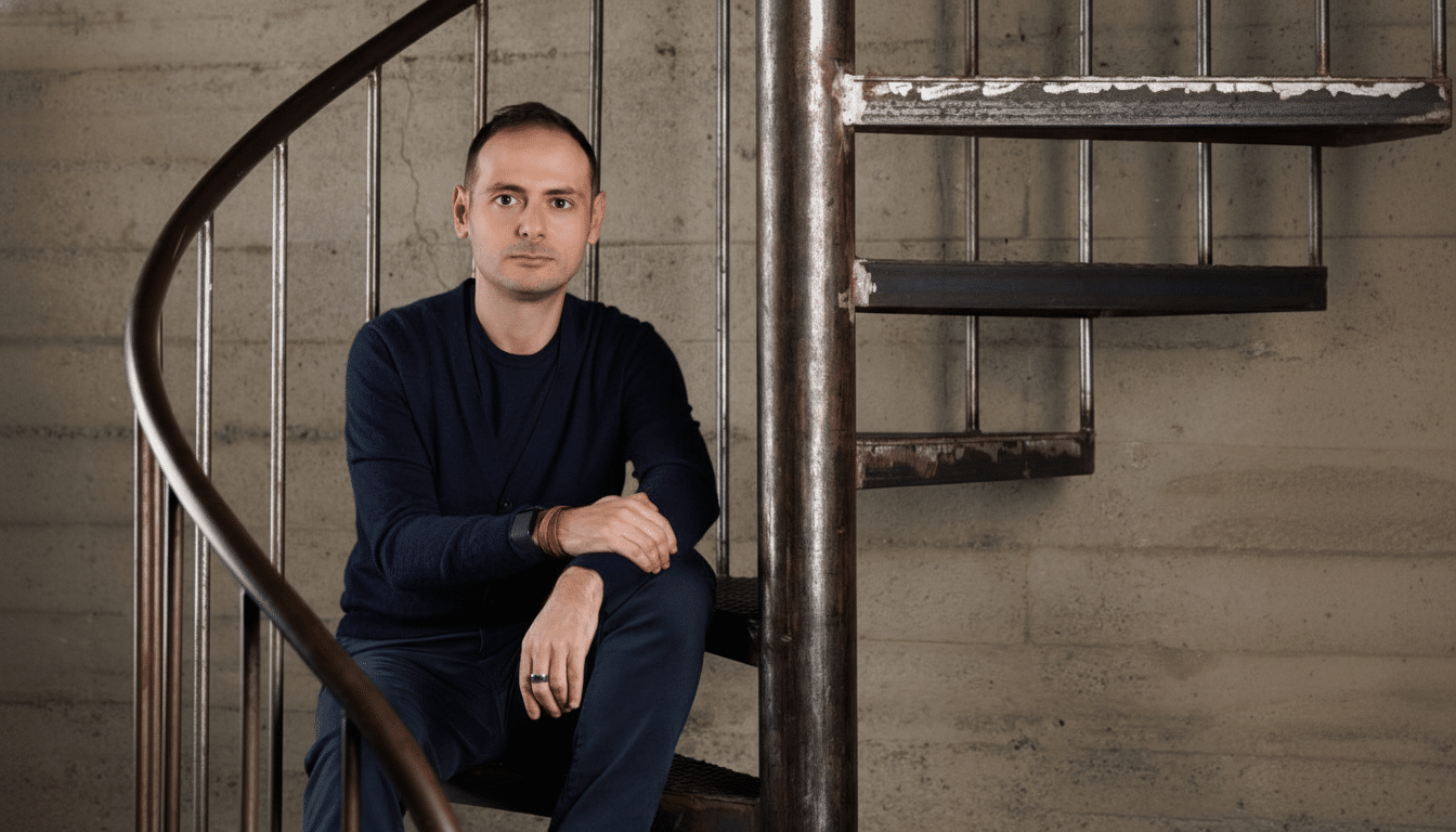 A man in a dark blue cardigan and pants sits on a spiral staircase, looking directly at the camera.
