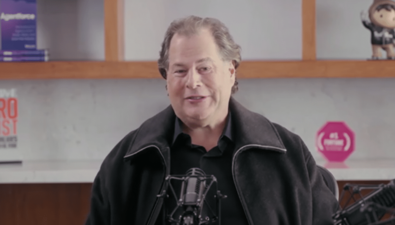 A man in a black jacket speaking into a microphone, with a blurred background of shelves and a small figurine.