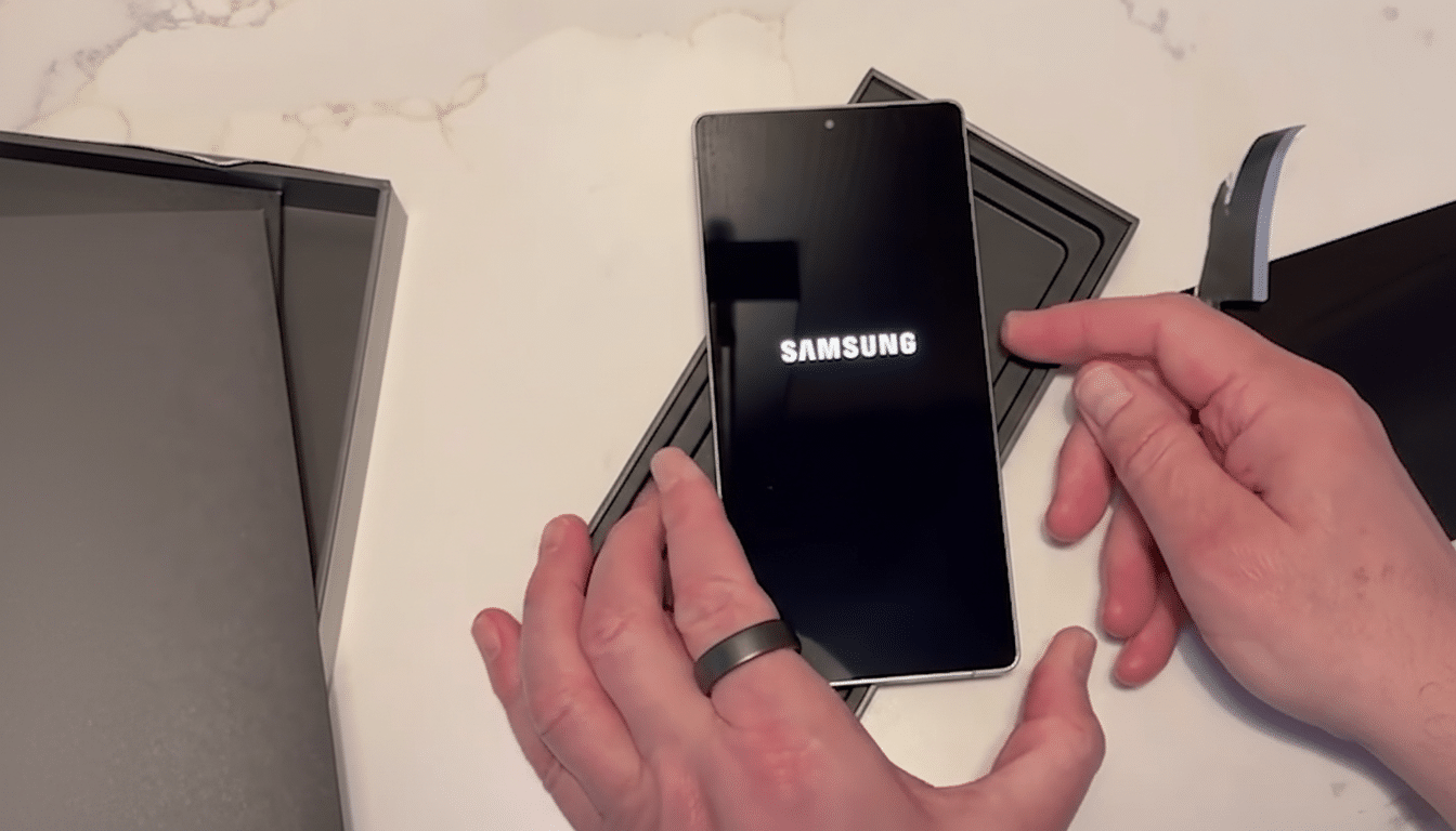 A persons hands holding a Samsung smartphone with the Samsung logo displayed on its screen, next to its packaging.