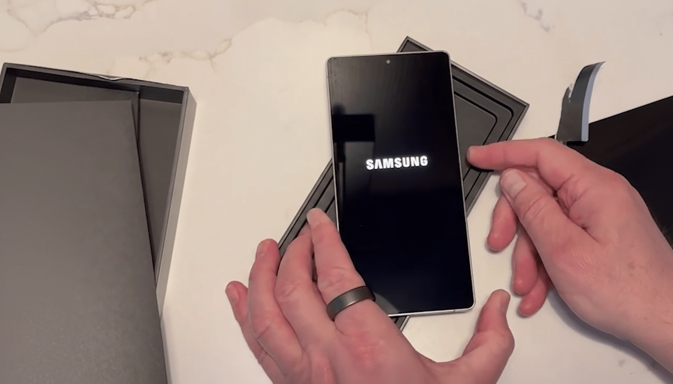 A persons hands holding a Samsung smartphone that is displaying the Samsung logo on its screen, with the phone partially in its packaging.