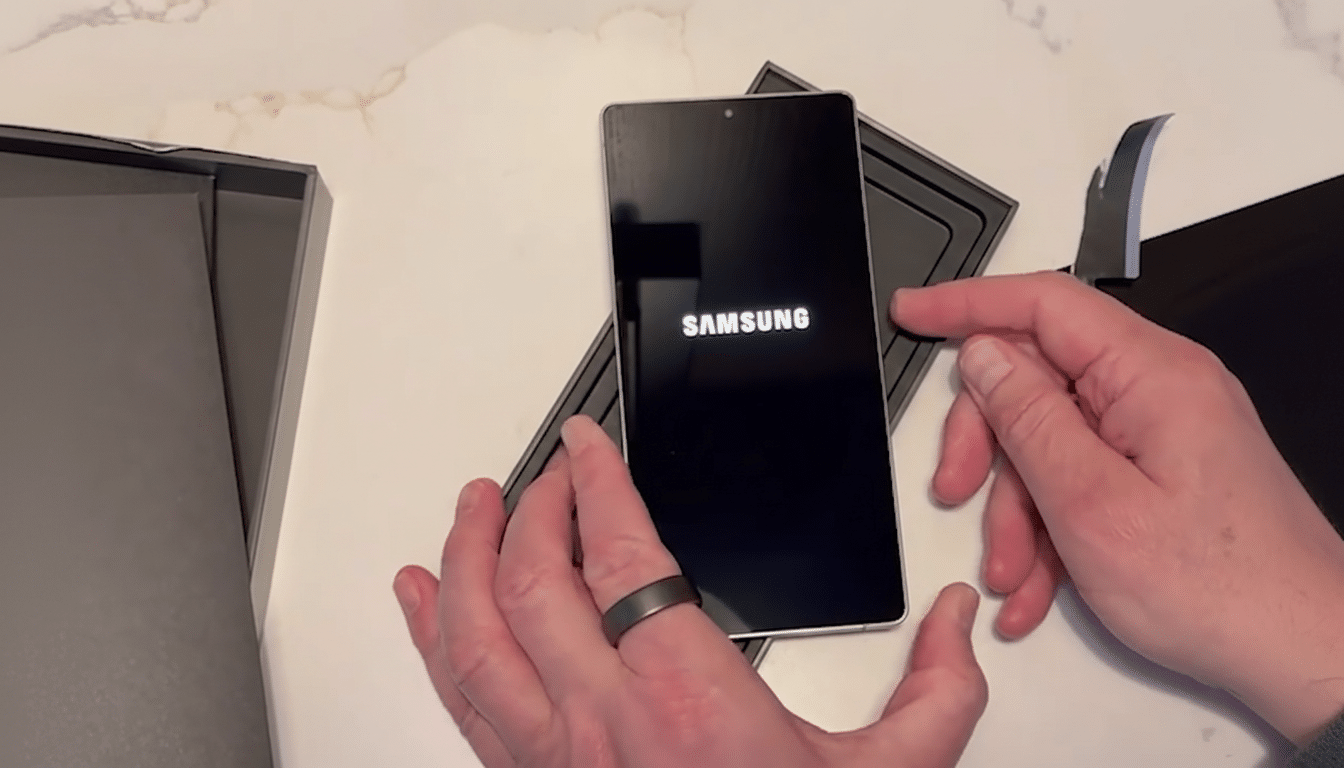 A persons hands holding a Samsung smartphone that is displaying the Samsung logo on its screen, with the phone partially in its box.