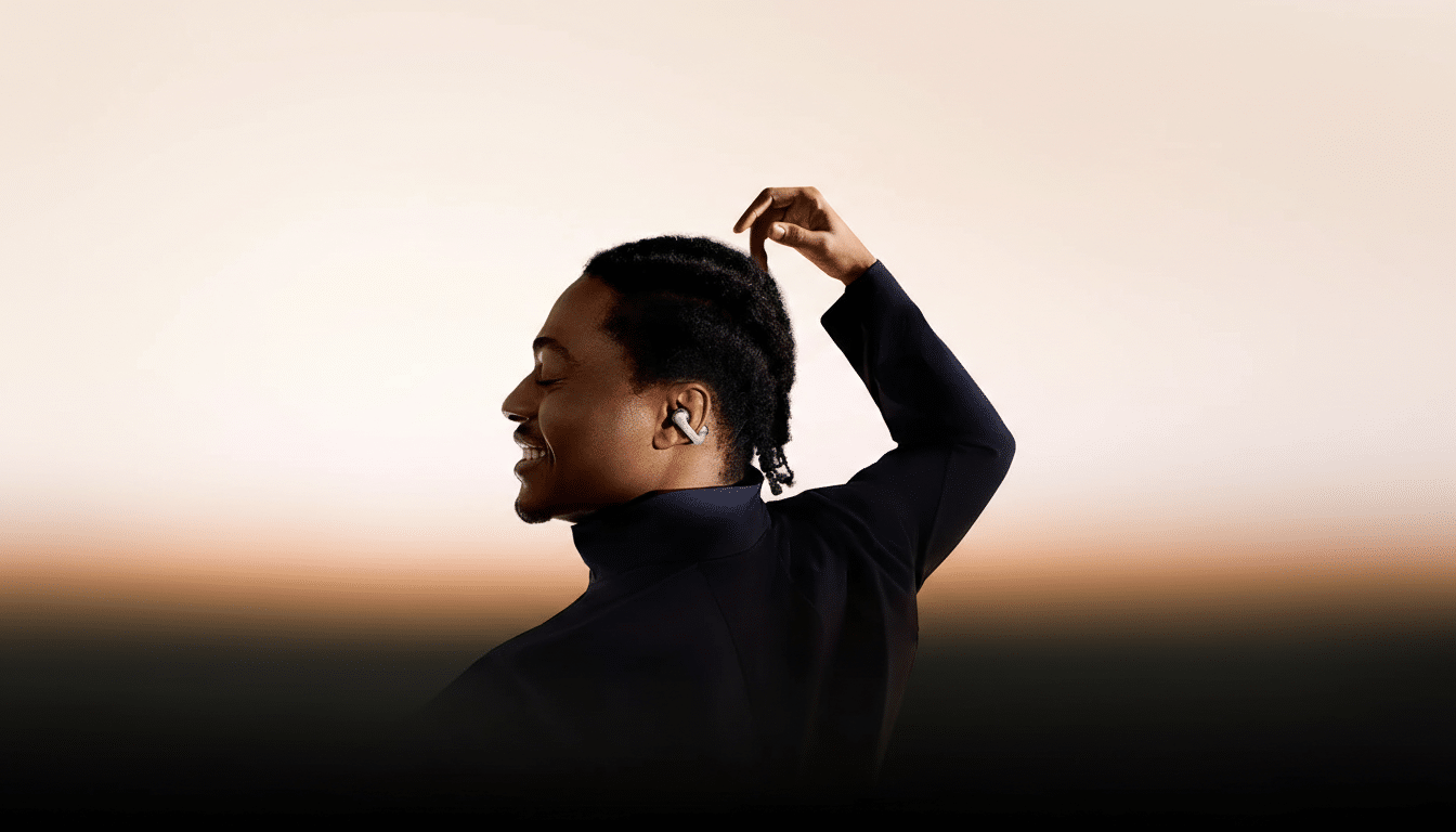 A man with an earbud in his ear, smiling and looking to the left, with his hand raised to his head.