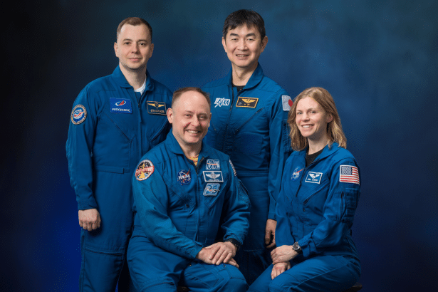Four astronauts in blue flight suits, two standing and two seated, smiling against a dark blue background.