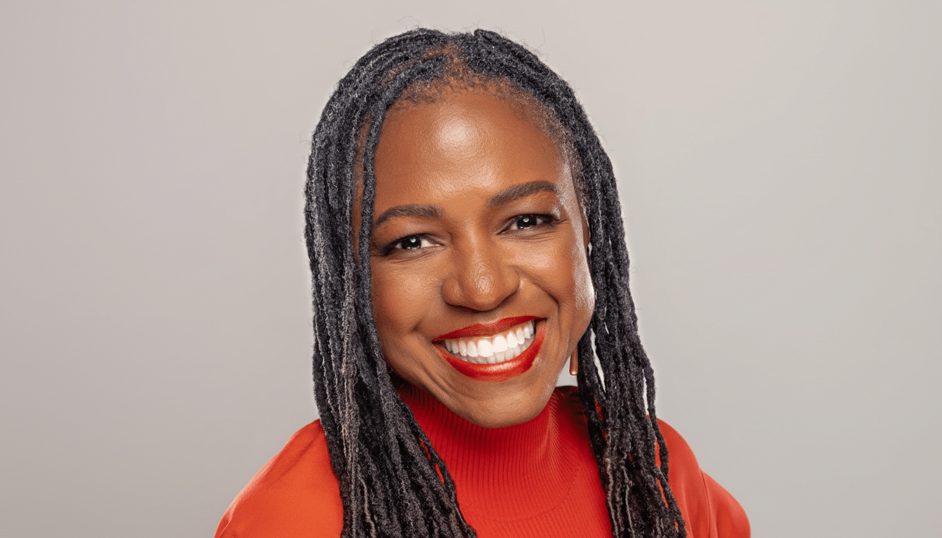 A professional headshot of a smiling woman with dark dreadlocks, wearing a red turtleneck, against a light gray background.