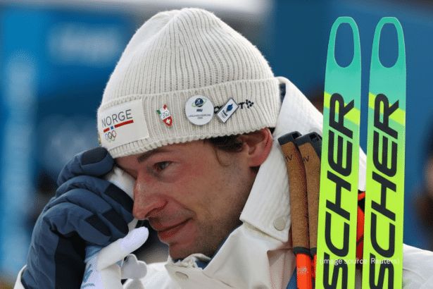 A close-up of a male skier in a white knit hat and jacket, wiping his eye with a gloved hand, with two green skis visible on the right side of the frame.