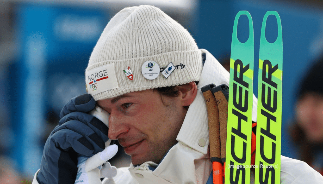 A close-up of a male skier in a white knit hat and jacket, wiping his eye with a gloved hand, with two green skis visible on the right side of the frame.