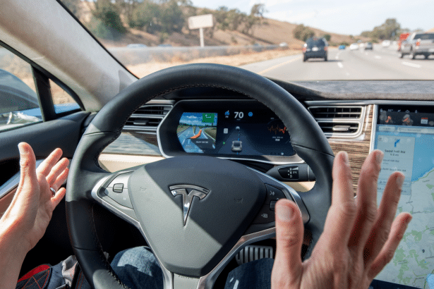 A persons hands are shown near the steering wheel of a Tesla car, which is driving on a highway. The cars dashboard displays navigation and speed information.