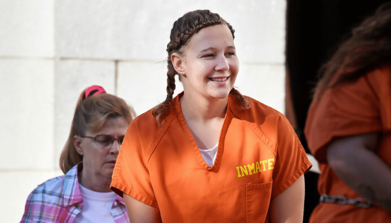 A young woman with braided hair, wearing an orange inmate uniform, smiles while looking to her right. Another woman with glasses and a plaid shirt is partially visible in the background.