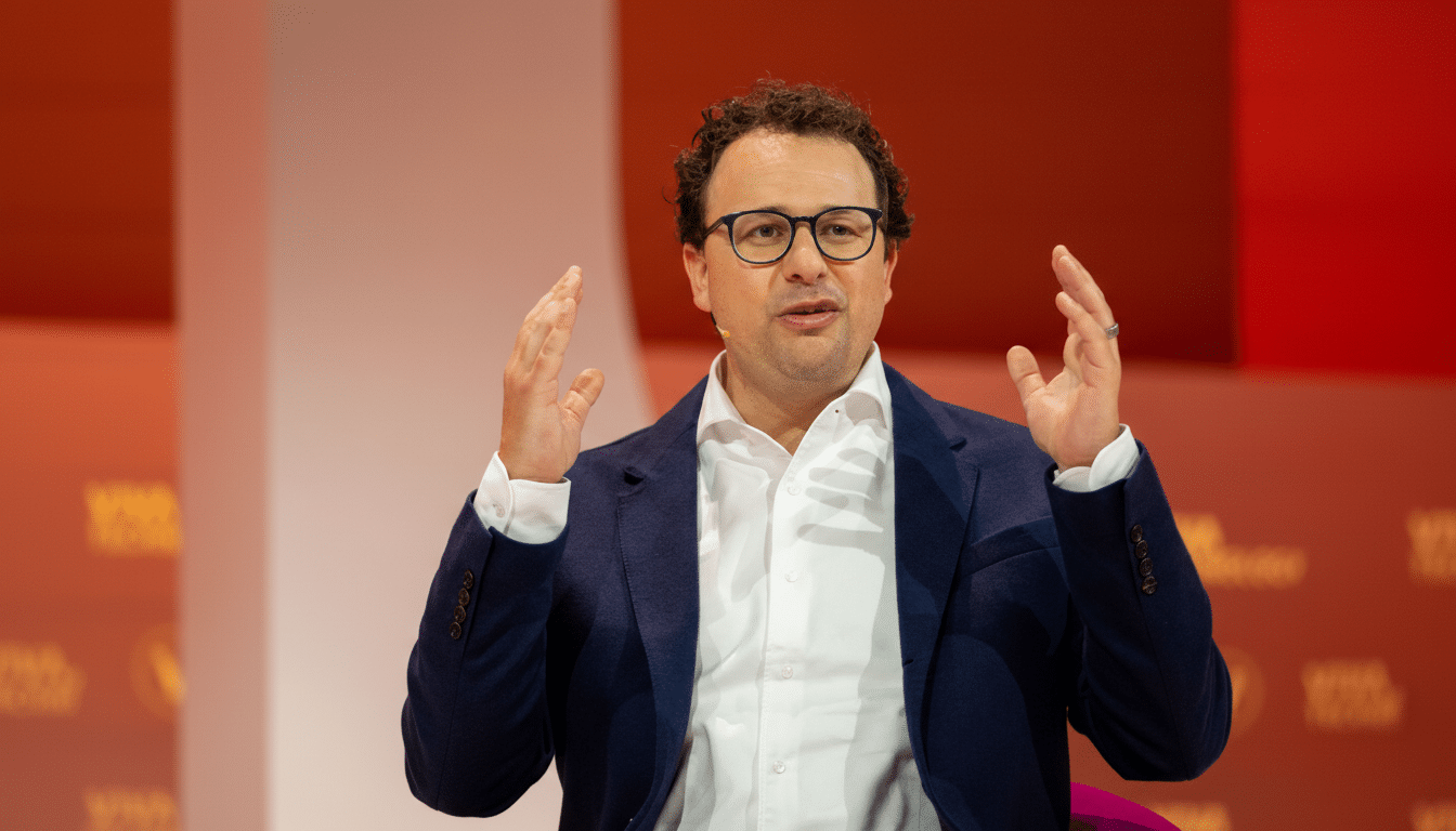 A man with curly dark hair and glasses, wearing a white shirt and a dark blue blazer, speaking with his hands raised in front of a red and white background.