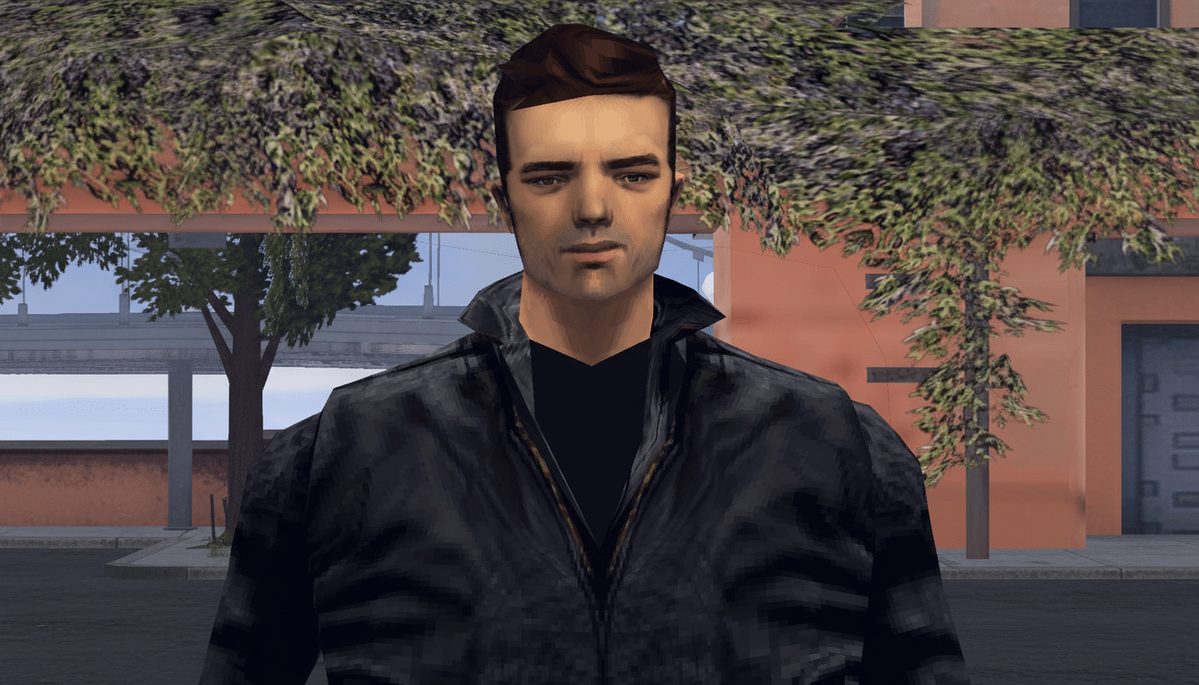 A man with dark hair and a black jacket stands in front of a building with trees and foliage.