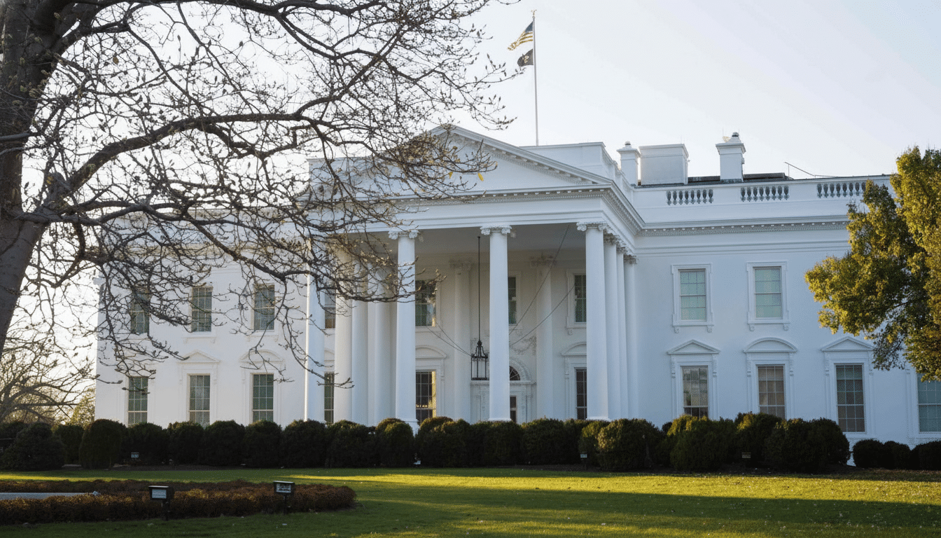 The White House, a large white building with columns, is seen from a distance with a tree in the foreground and a flag flying above.
