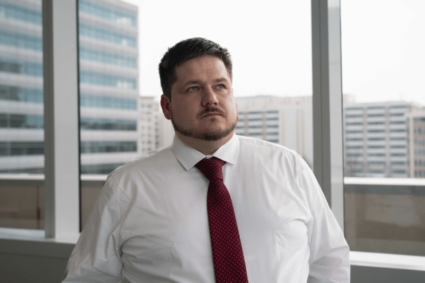A man in a white shirt and red tie looking up and to the right, with a city building visible through a window in the background.