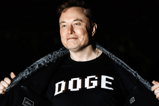 Elon Musk wearing a black t-shirt with DOGE written on it, opening his jacket.