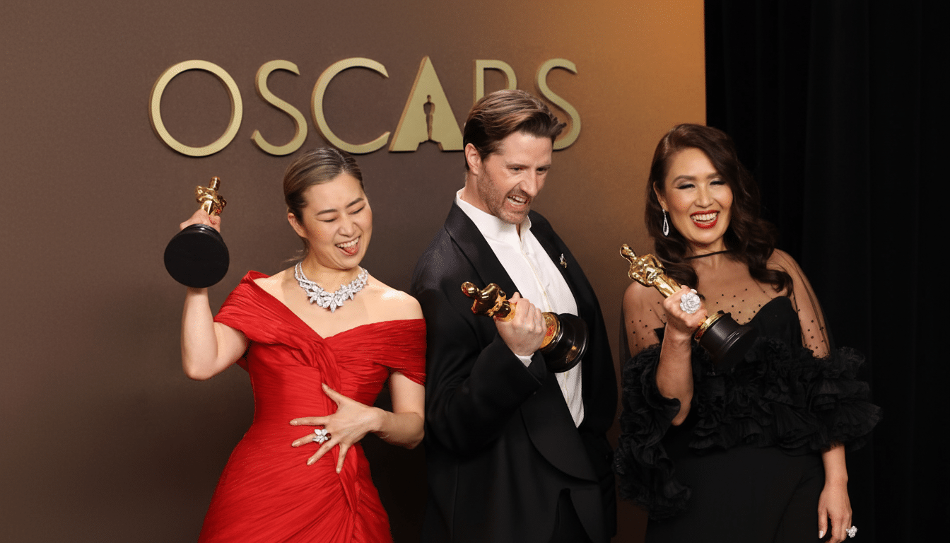 Three people, two women and one man, are holding Oscar awards and making playful, excited faces in front of a backdrop that says OSCARS. The woman on the left is wearing a red off-the-shoulder dress and a large diamond necklace, sticking her tongue out. The man in the center is wearing a tuxedo and has a wide, joyful expression. The woman on the right is wearing a black dress with sheer sleeves and ruffles, smiling broadly.
