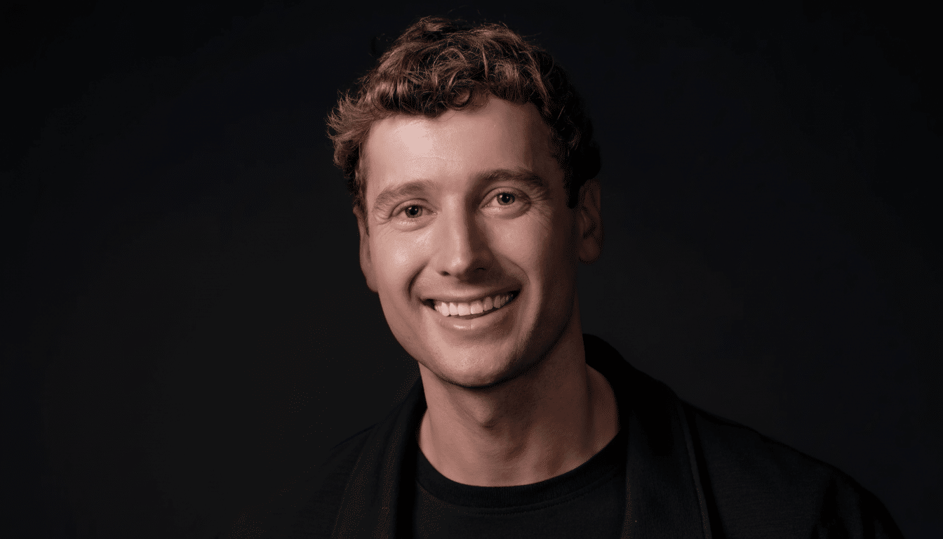 A professional headshot of a smiling man with curly brown hair and blue eyes, wearing a black t-shirt and jacket, against a dark gray background.