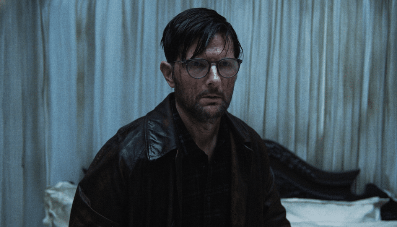 A man with dark hair, a beard, and glasses, wearing a dark leather jacket and shirt, looking forward with a serious expression against a muted, light-colored background.