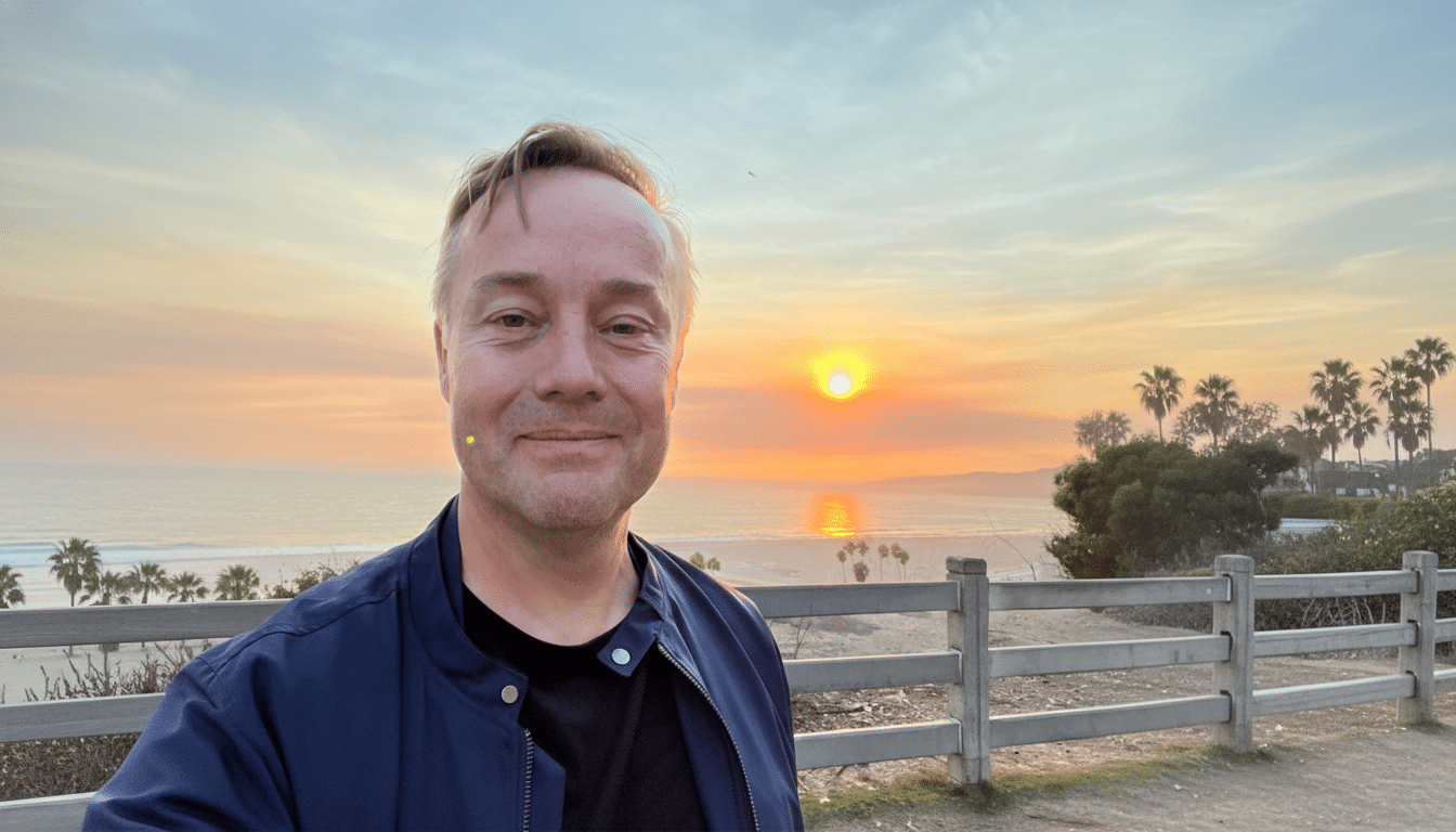 A man smiling at the camera with a sunset over the ocean in the background.