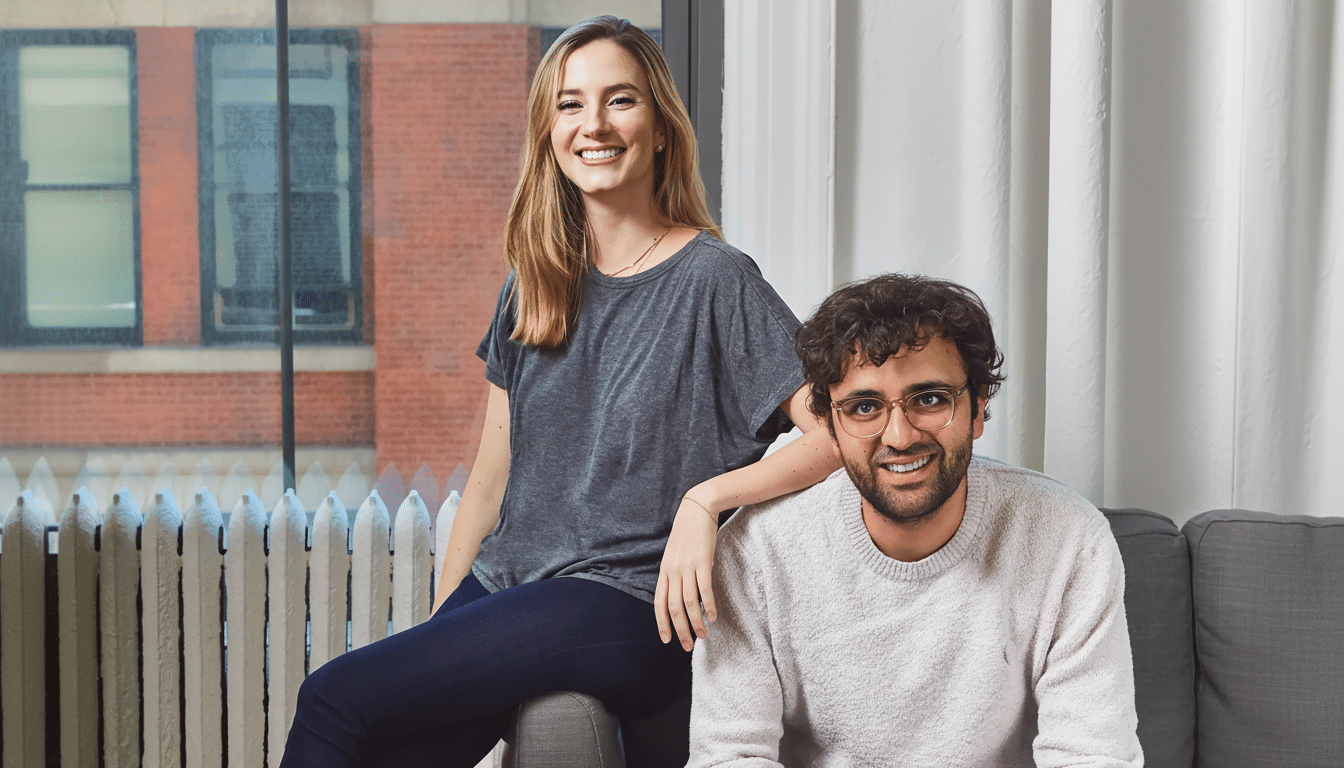 A man and a woman smiling at the camera, with the woman sitting on a ledge and the man sitting on a couch.