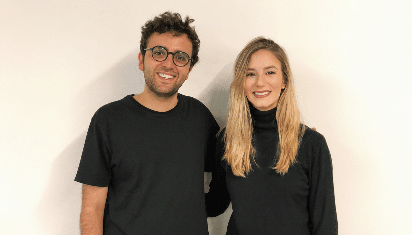 A man and a woman smiling, both wearing black tops, standing against a white background.