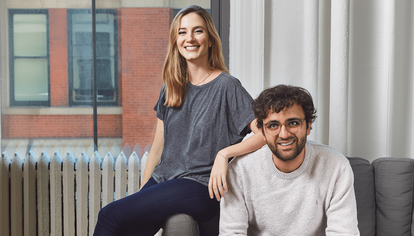 A smiling woman in a grey shirt and dark pants sits on a grey couch, leaning against a man in a light sweater and glasses who is also smiling and seated on the couch. They are in a room with a large window showing a brick building outside.