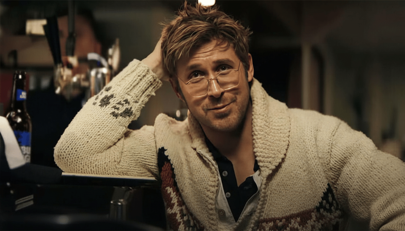 A man with messy blonde hair and glasses, wearing a cream-colored cardigan, leans on a bar with a slight smile.