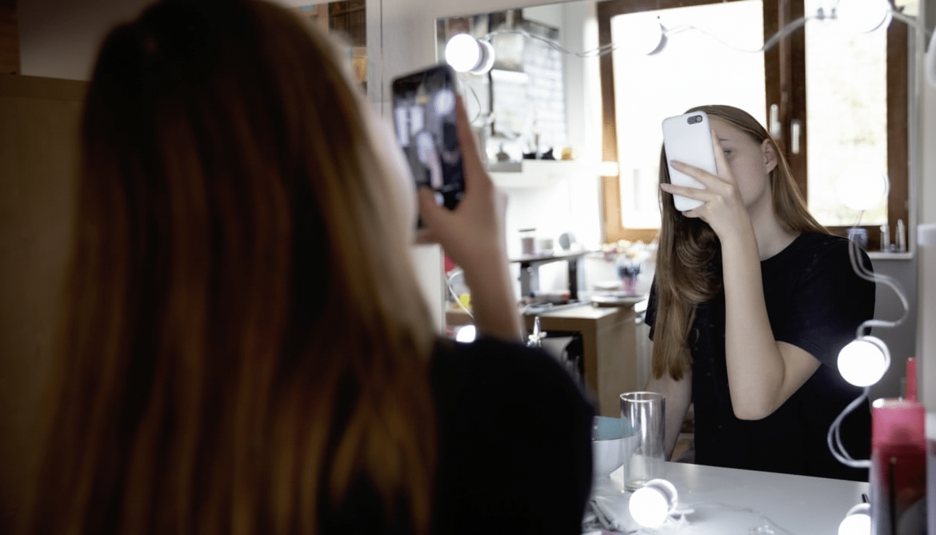 A young woman taking a selfie in a mirror, holding a white smartphone to her face.