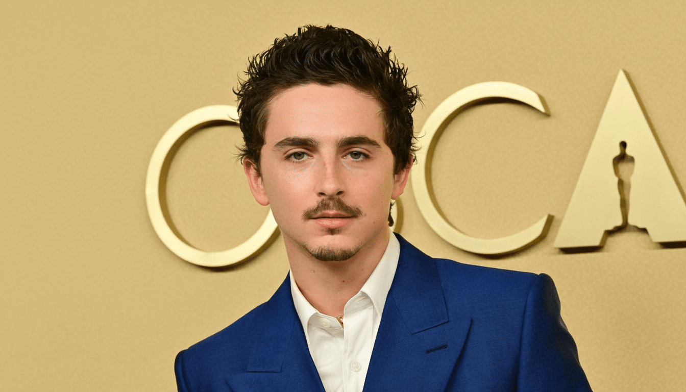 A young man with dark, curly hair and a mustache, wearing a blue suit and white shirt, stands in front of a gold background with the word OSCARS partially visible.
