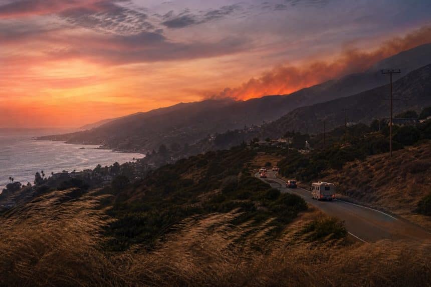 Wind-bent dry grass on a Malibu hillside with distant wildfire glow and vehicles leaving on a canyon road.